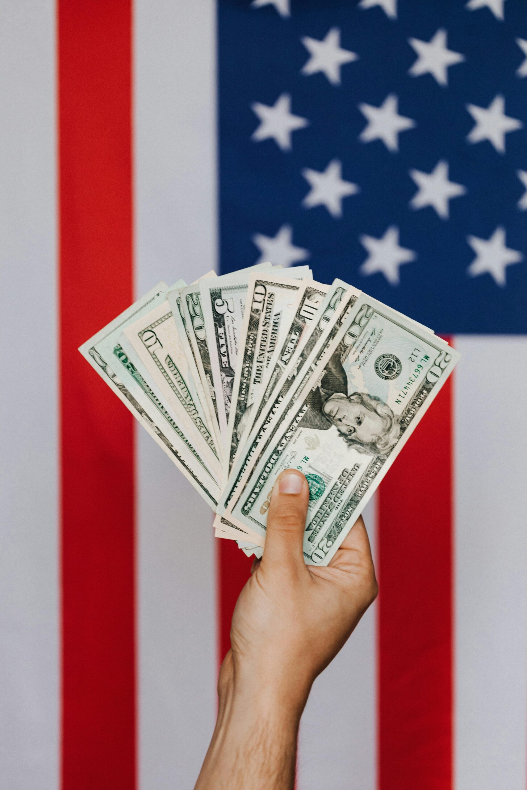 A hand holding US dollar bills in front of an American flag, symbolizing wealth and patriotism.